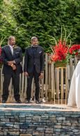 Two men in dark suits standing outdoors beside red flowers and a wooden fence