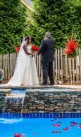 Wedding couple standing by a pool, exchanging vows in a garden with a floral arch and stone waterfall backdrop