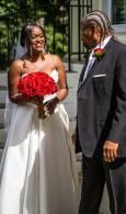 Bride in white dress holds red bouquet beside groom in dark suit outdoors