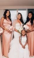 Four people in matching blush dresses and a white gown, posing together indoors with bouquets.
