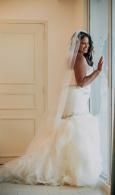 Bride in a white wedding dress standing by a window, looking out with a veil and train flowing behind her
