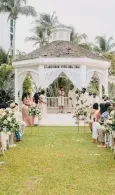 Outdoor wedding gazebo decorated with white drapes and flowers, lining a grassy aisle at a beachside venue