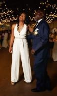 Two people in formal attire stand together under warm string lights, smiling at an evening event.