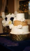 Three-tier white wedding cake with gold accents and white flowers on a dark table.