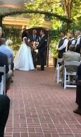 Bride and groom standing at an outdoor wedding ceremony under a canopy, guests seated along a brick aisle.