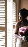 Bride holding a pink bouquet beside sunlit shutters, wearing a white dress.