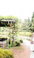Garden walkway with lush greenery, a white pergola, and a person standing at the right edge