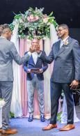 Man under floral arch receives certificate from two suited men in a formal ceremony