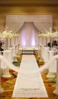 Elegant wedding aisle with white chairs, floral arrangements, and a draped stage backdrop in warm lighting