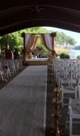 Covered outdoor wedding aisle with white chairs, floral decorations, and a draped altar by the water