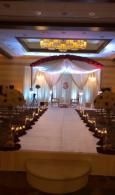 Decorated wedding hall with a central aisle, white drapery, chandeliers, and rows of chairs