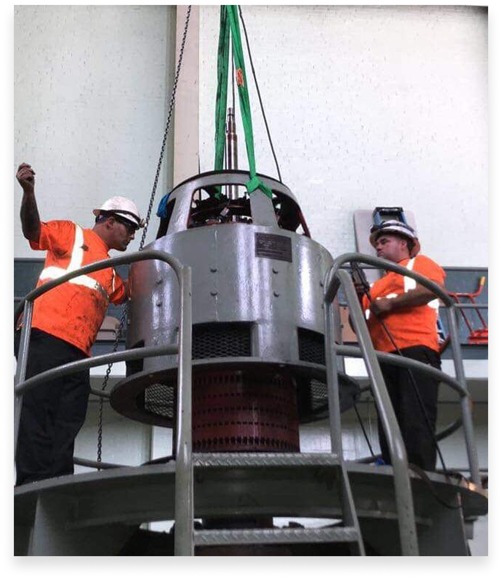 Two workers in orange vests oversee the lifting of a large machine in a facility.