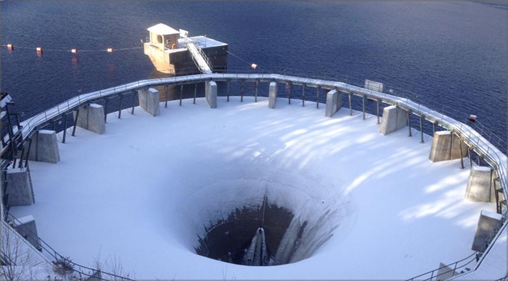 Snow-covered dam outlet with a deep circular opening. A boat is moored nearby on the water.