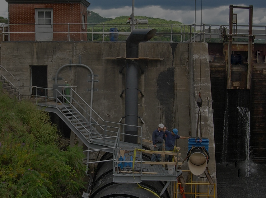 Workers on scaffolding near a dam, examining a large pipe. Building and water in background.