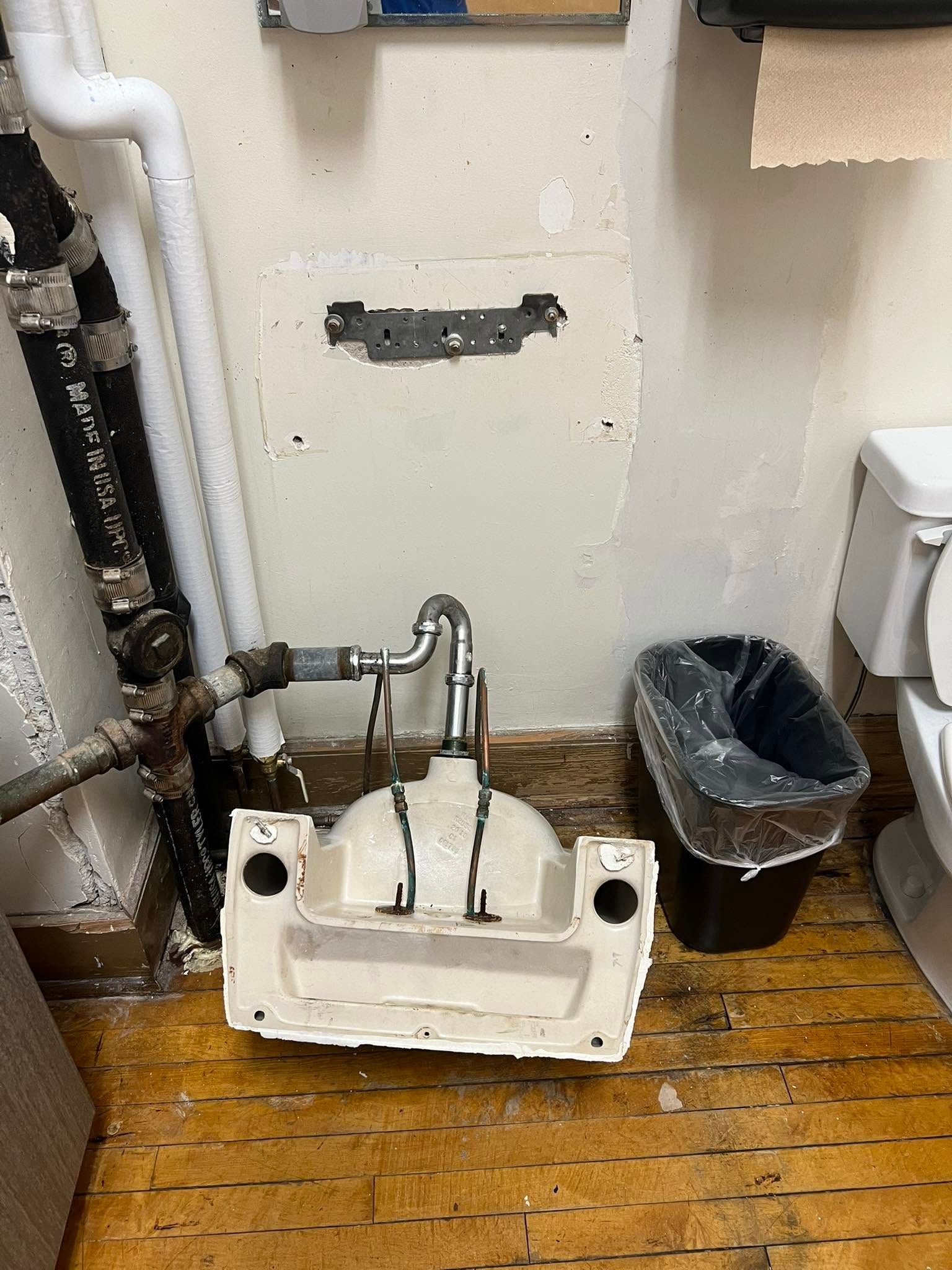 Detached sink basin and exposed pipes on a wood floor in a utility room, with wall damage behind it