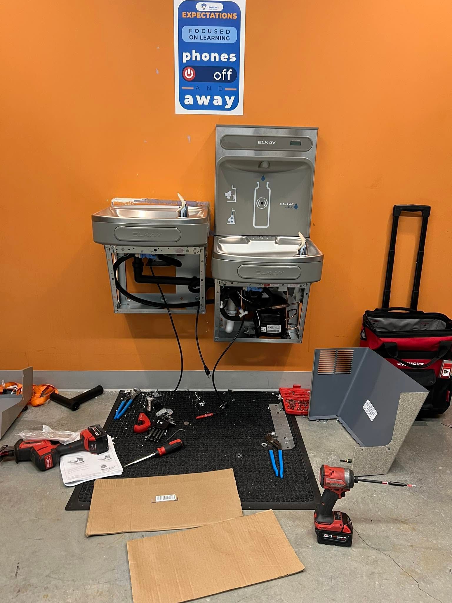 Workshop wall with a blue phone sign, utility sinks, tools on the floor, and an open laptop on the right.