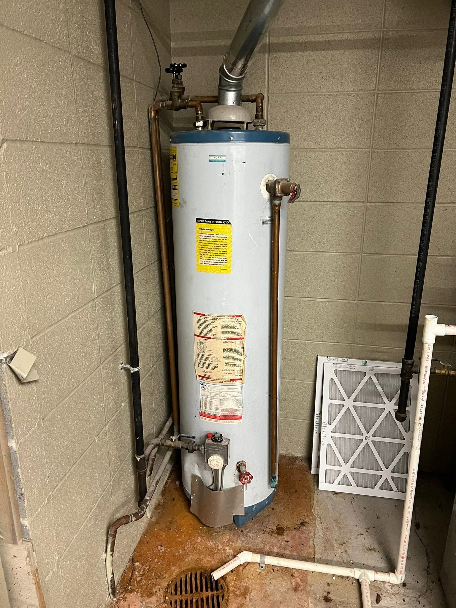White water heater in a narrow utility closet with exposed pipes and a floor drain.