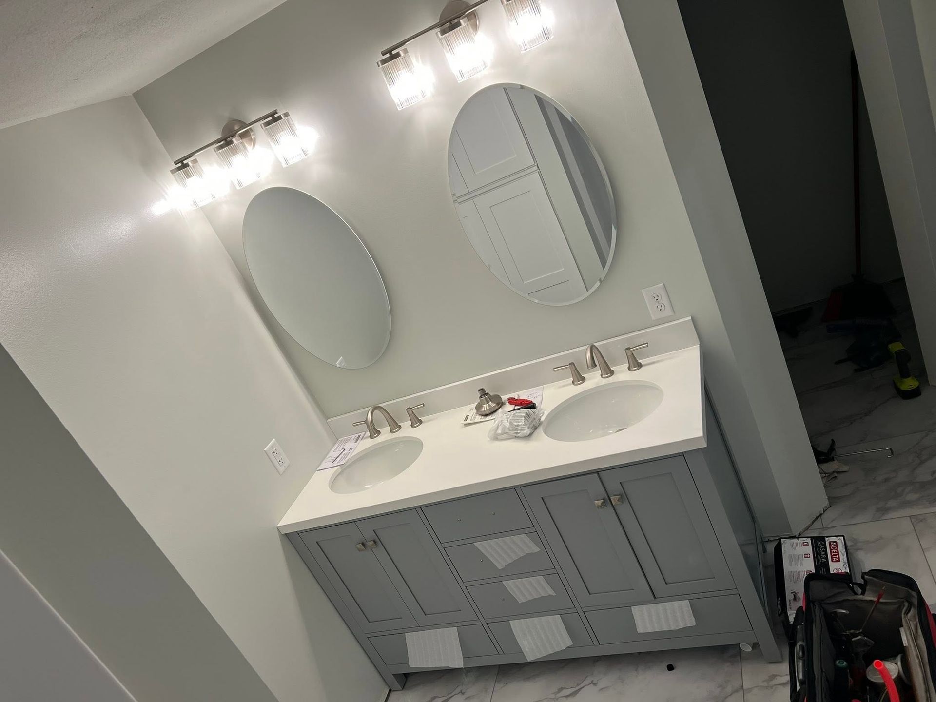 Bathroom vanity with double sinks, oval mirrors, and bright wall lights; cleaning supplies on the counter.