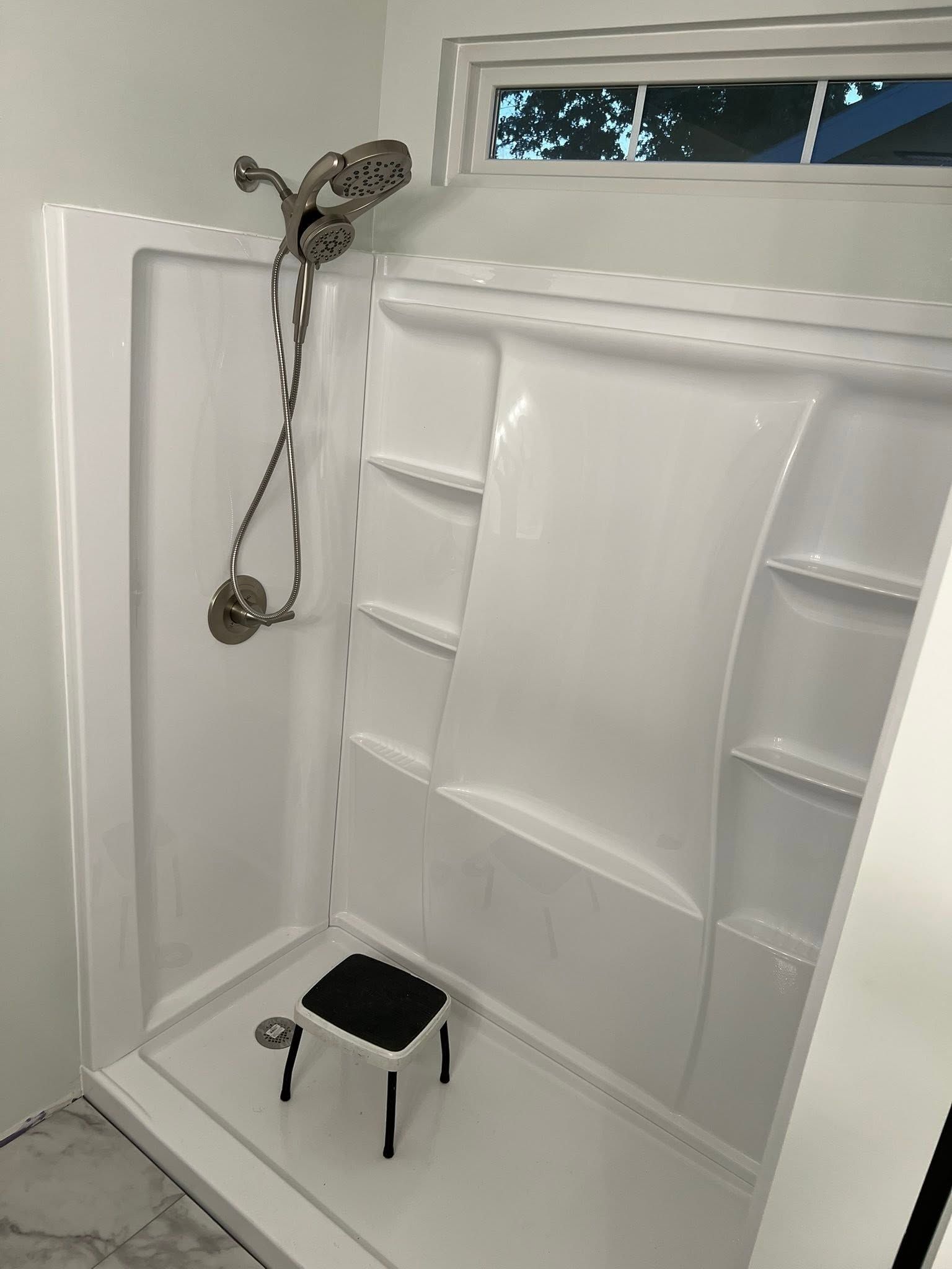 White bathroom shower with built-in shelves, grab bars, and a black shower stool near the drain.