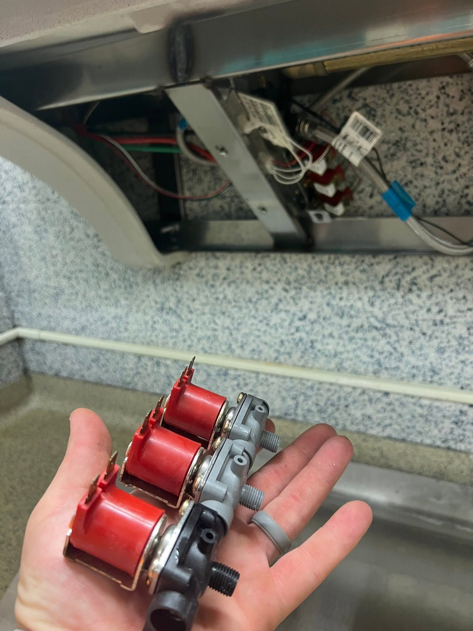 Hand holding a small red and silver mechanical part near exposed wiring inside a machine.