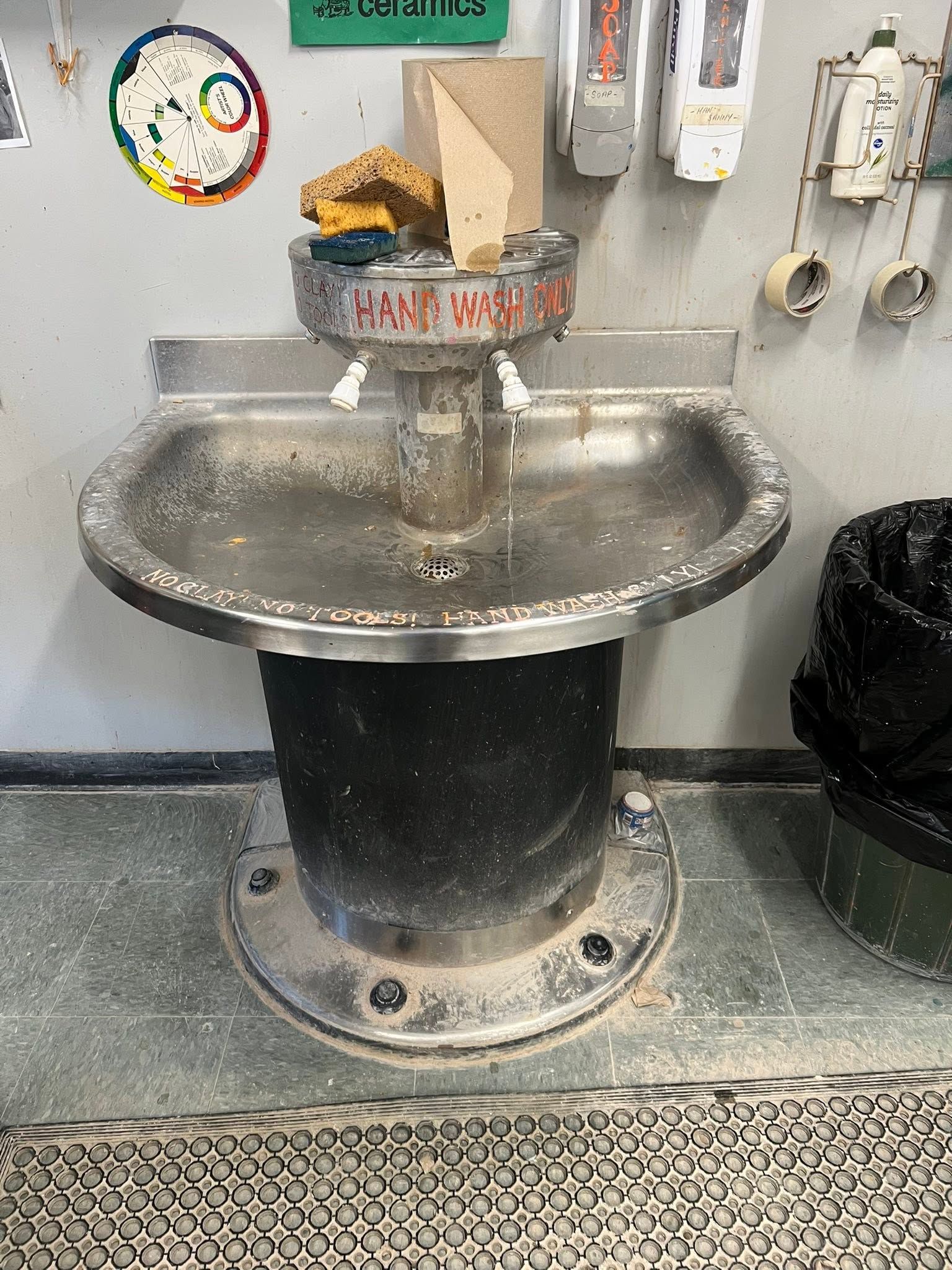 Public sink with metal basin, soap dispensers, and a trash can in a restroom or utility area