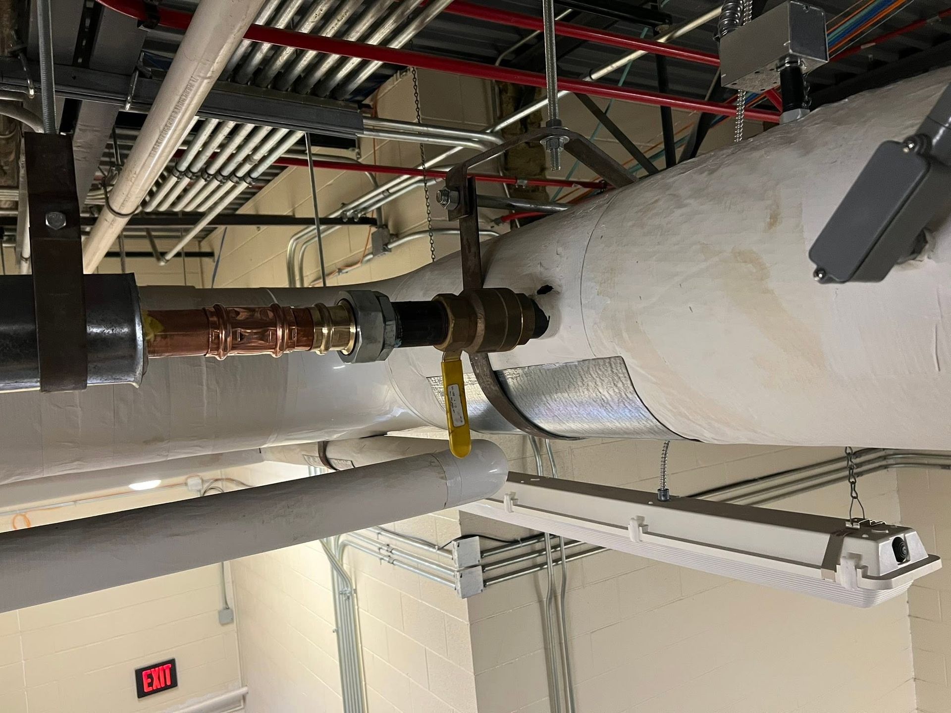 Industrial ceiling pipes and ductwork in a utility room, with a large insulated pipe and overhead cables.