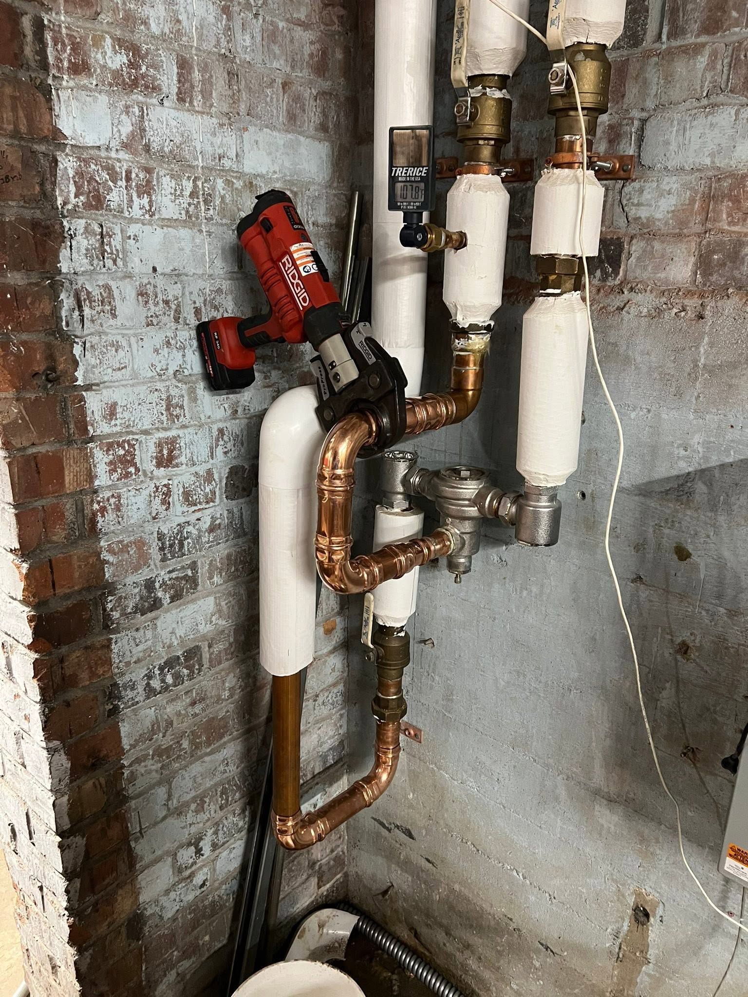 Plumbing valves and insulated pipes mounted on a rough brick wall in a basement-like utility area
