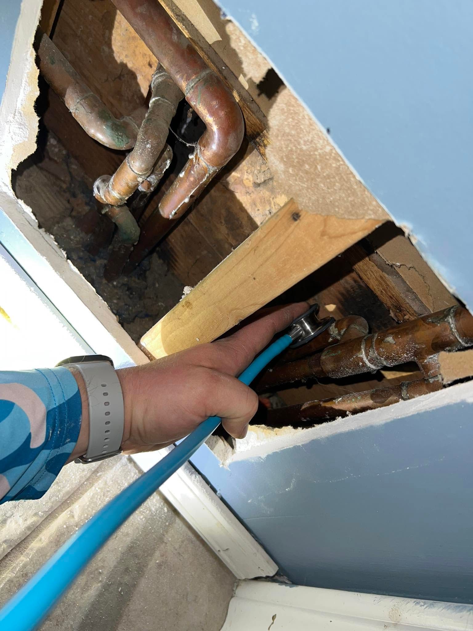 Hand reaching into a ceiling opening with blue tool hose near exposed pipes and wood framing
