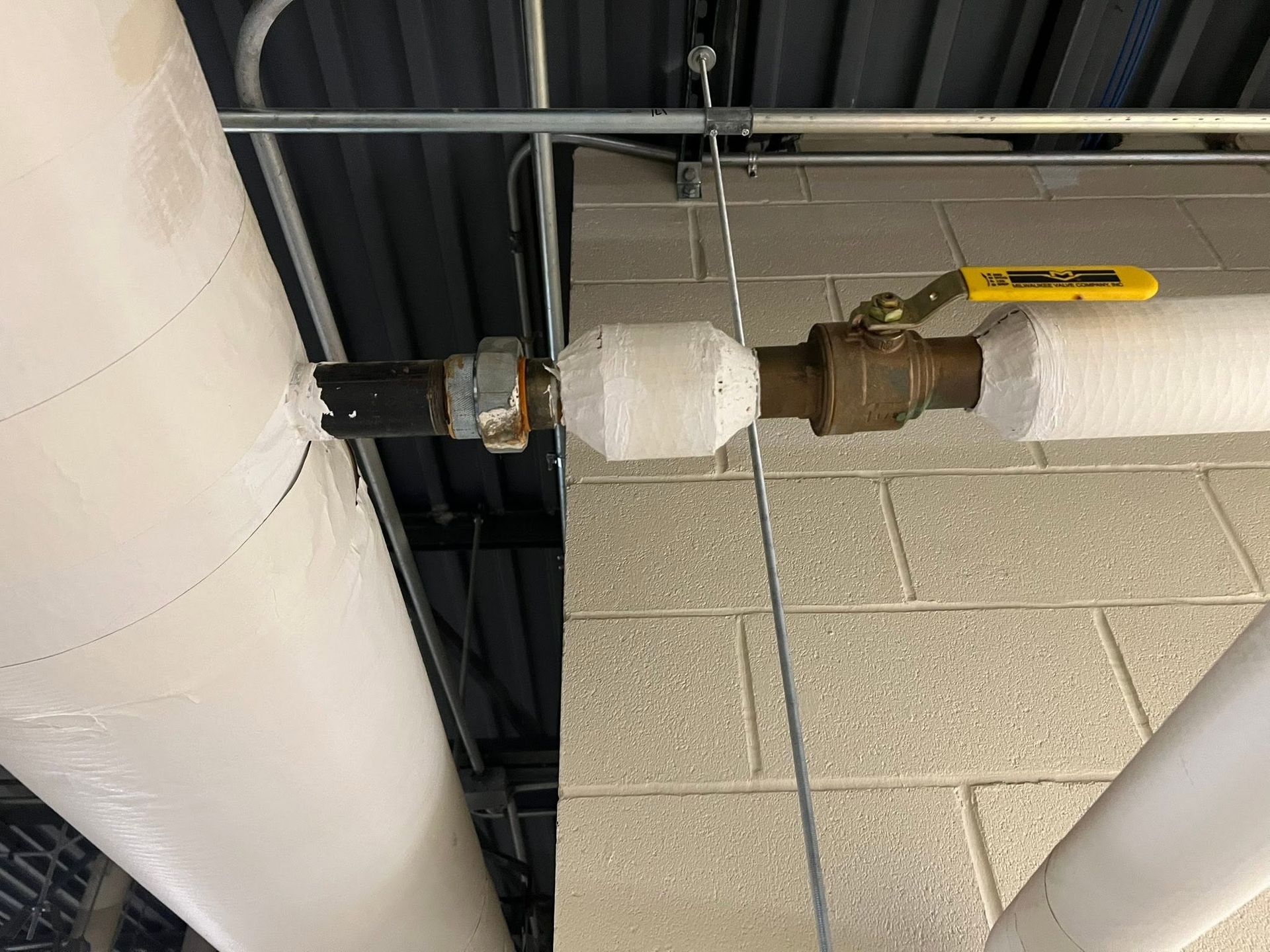 Exposed white pipe joint and valve near ceiling tiles and black ductwork in an industrial interior