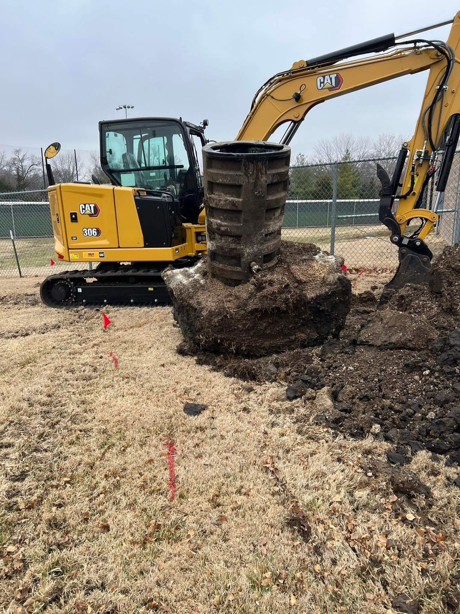 Yellow excavator lifting a tree root ball in a gravel lot under an overcast sky