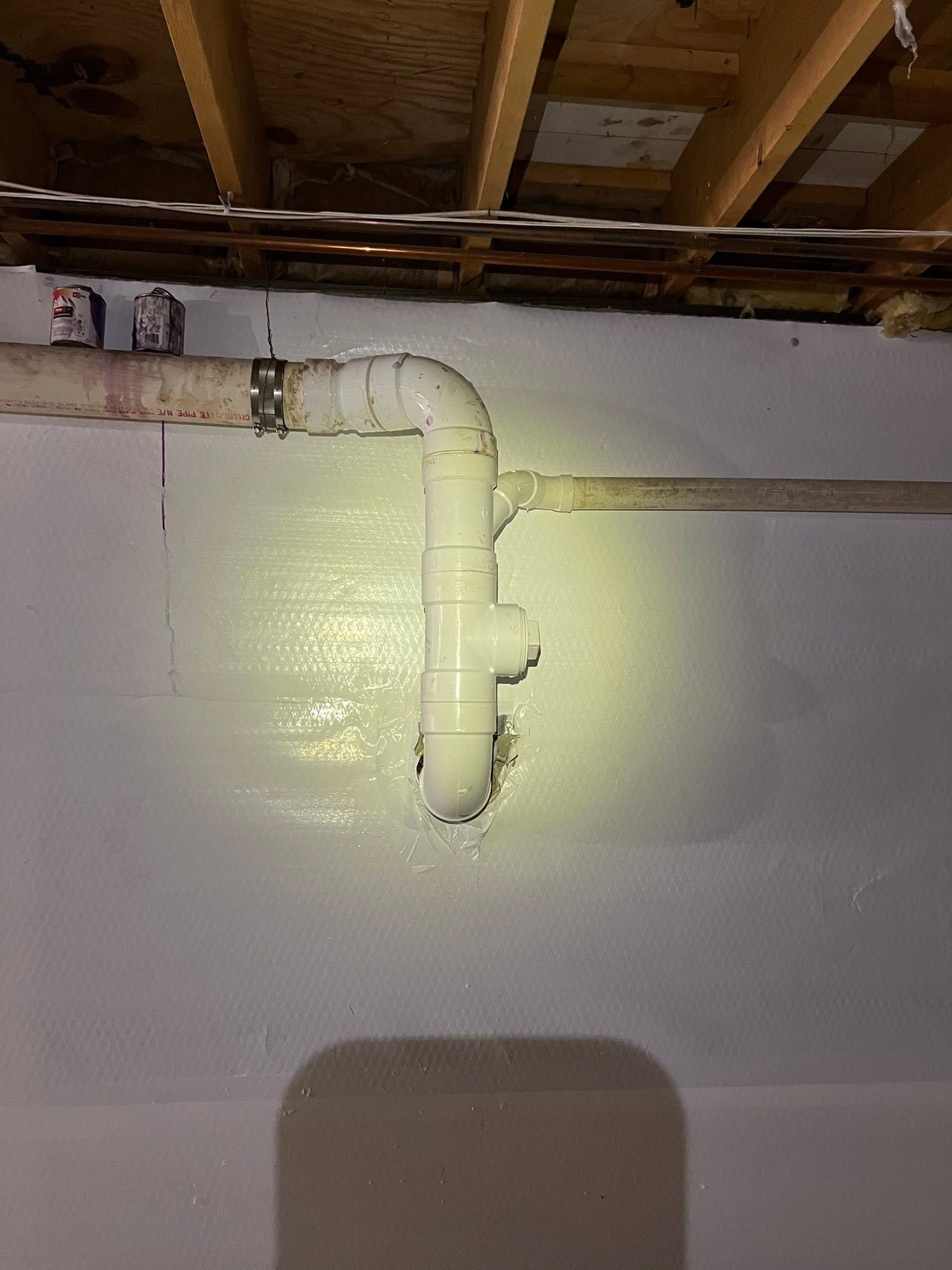 White-painted plumbing pipe with joints and a valve on a basement wall beneath exposed ceiling beams