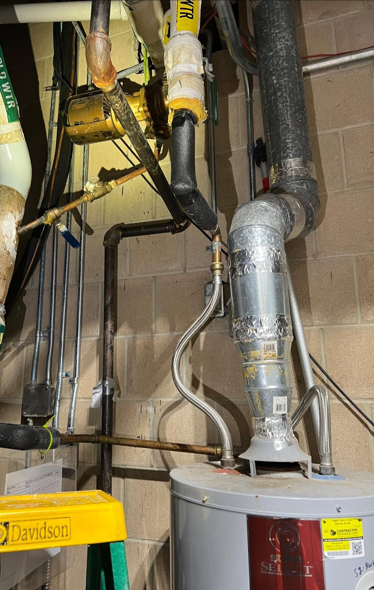 Ductwork and pipes above a water heater in a utility room, with yellow equipment and cinder block walls