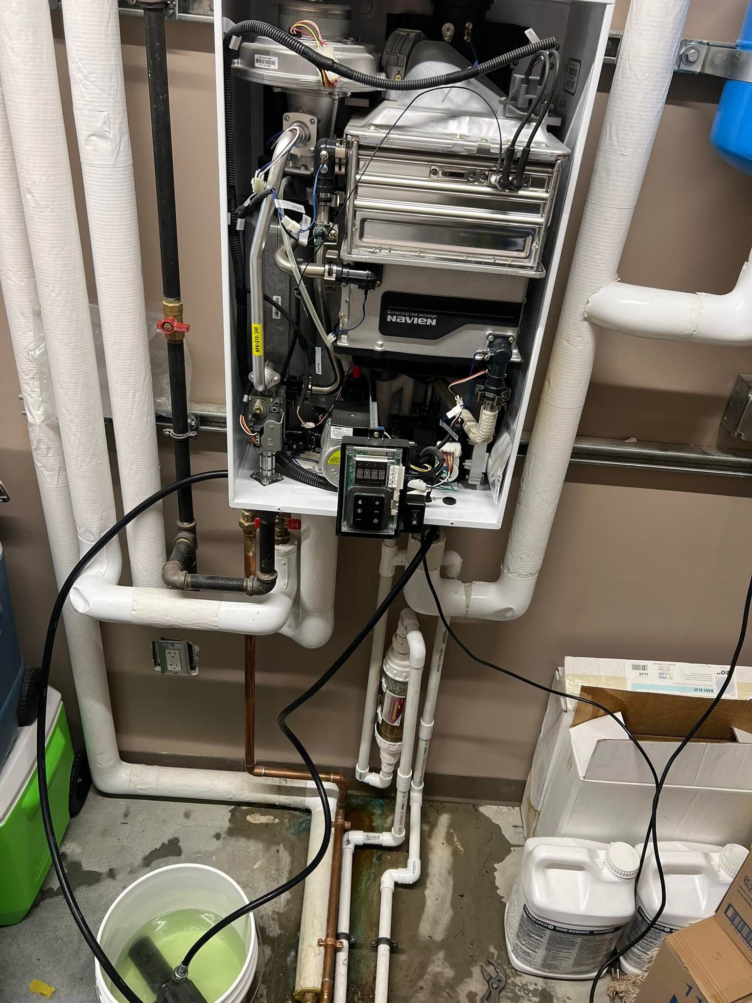 Open water heater and plumbing pipes in a utility closet, with buckets and wires on the floor.