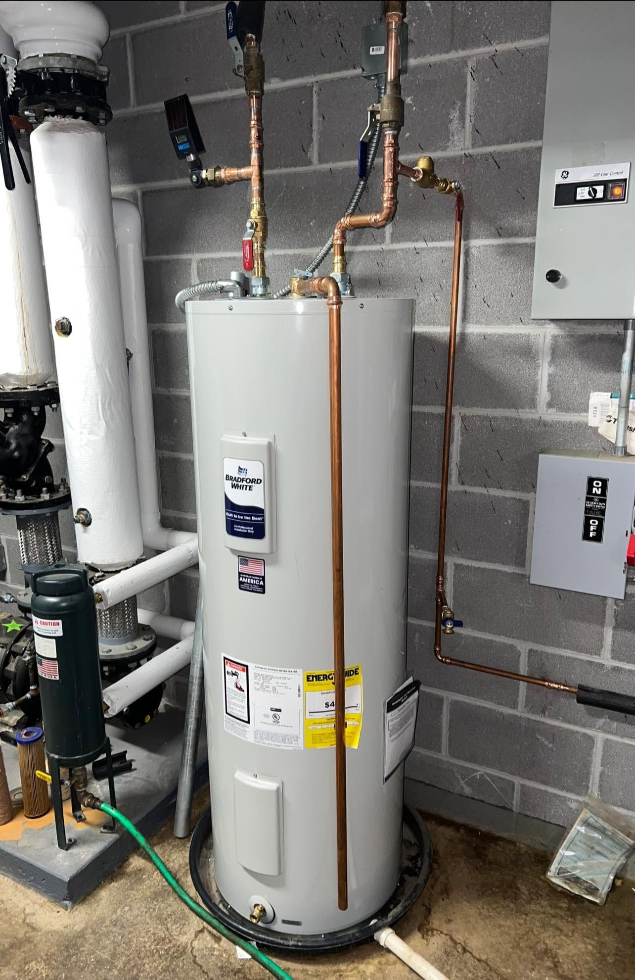 Water heater in a utility room with exposed copper pipes, gray cinderblock walls, and nearby electrical boxes