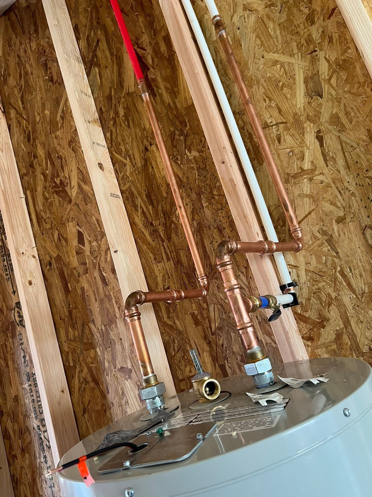 Copper pipes and fittings above a white water heater against unfinished plywood walls.
