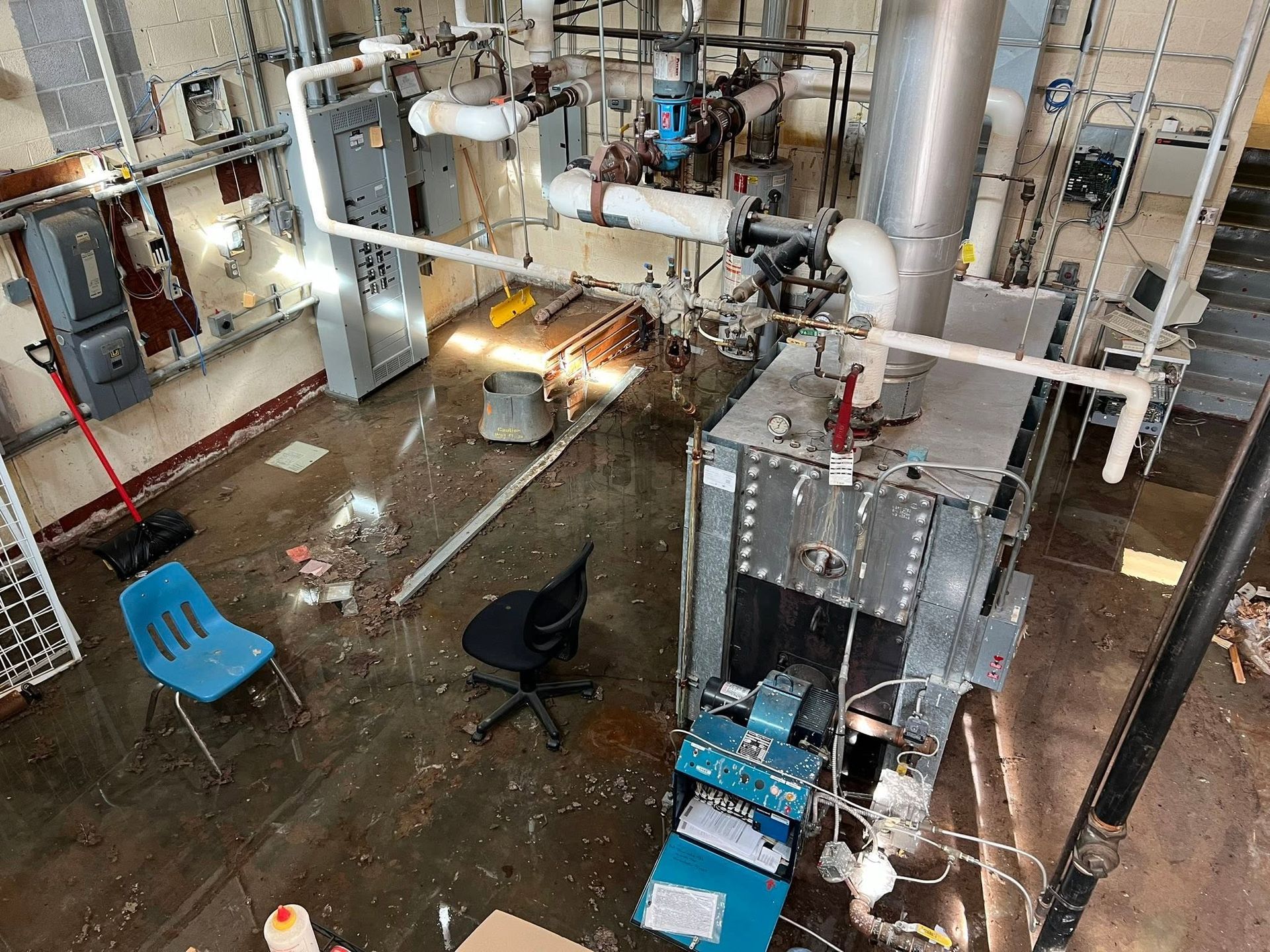 Industrial facility interior with large metal piping, wet floor, and scattered blue chairs