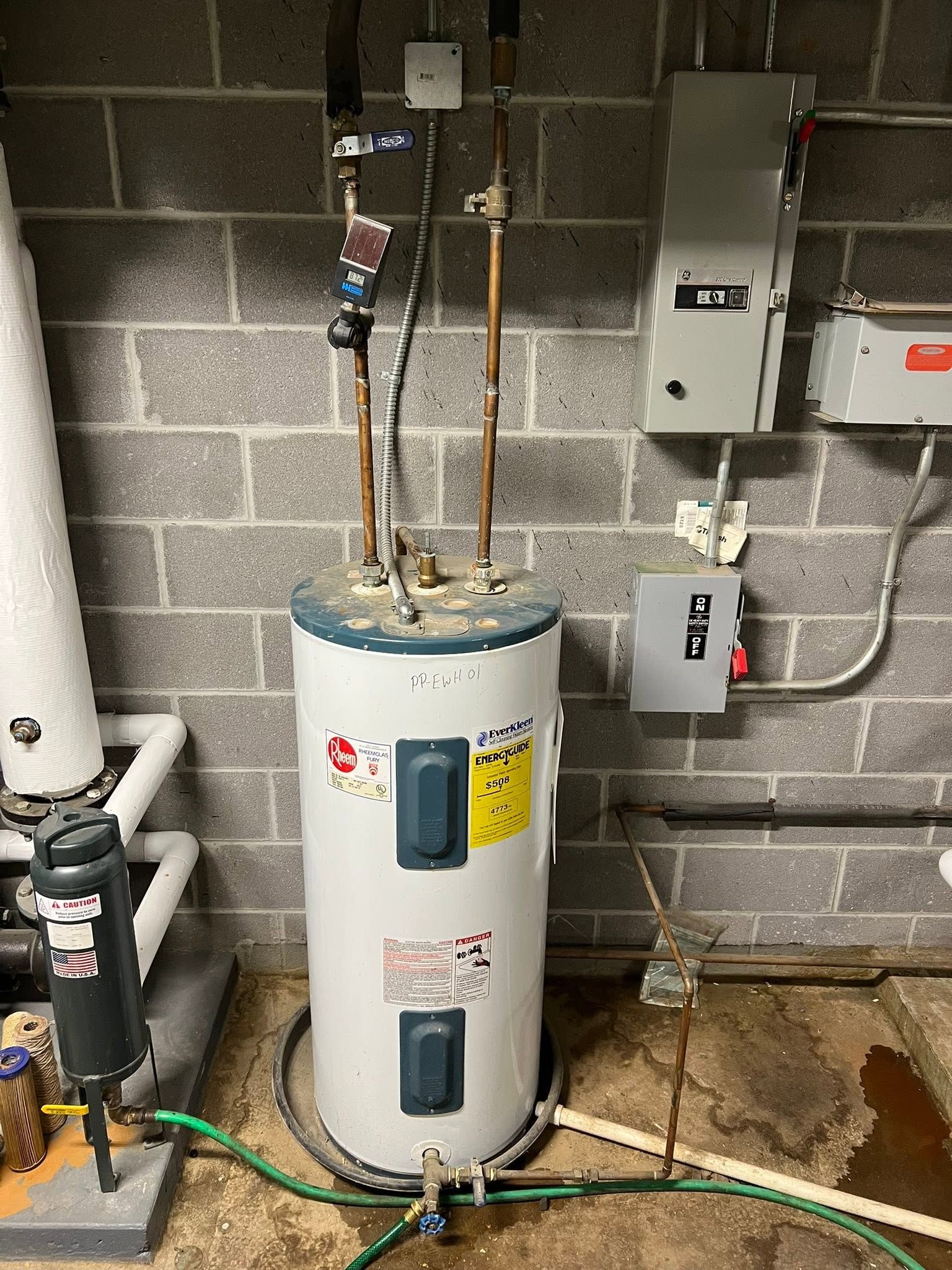 Water heater in a basement utility room against cinder block wall with exposed pipes and electrical boxes