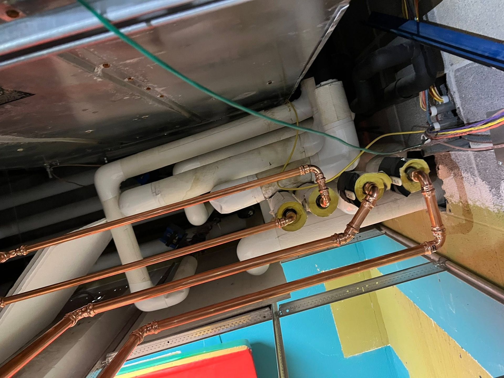 Underside of a table with exposed pipes, wiring, and yellow brackets against a blue-and-yellow wall.
