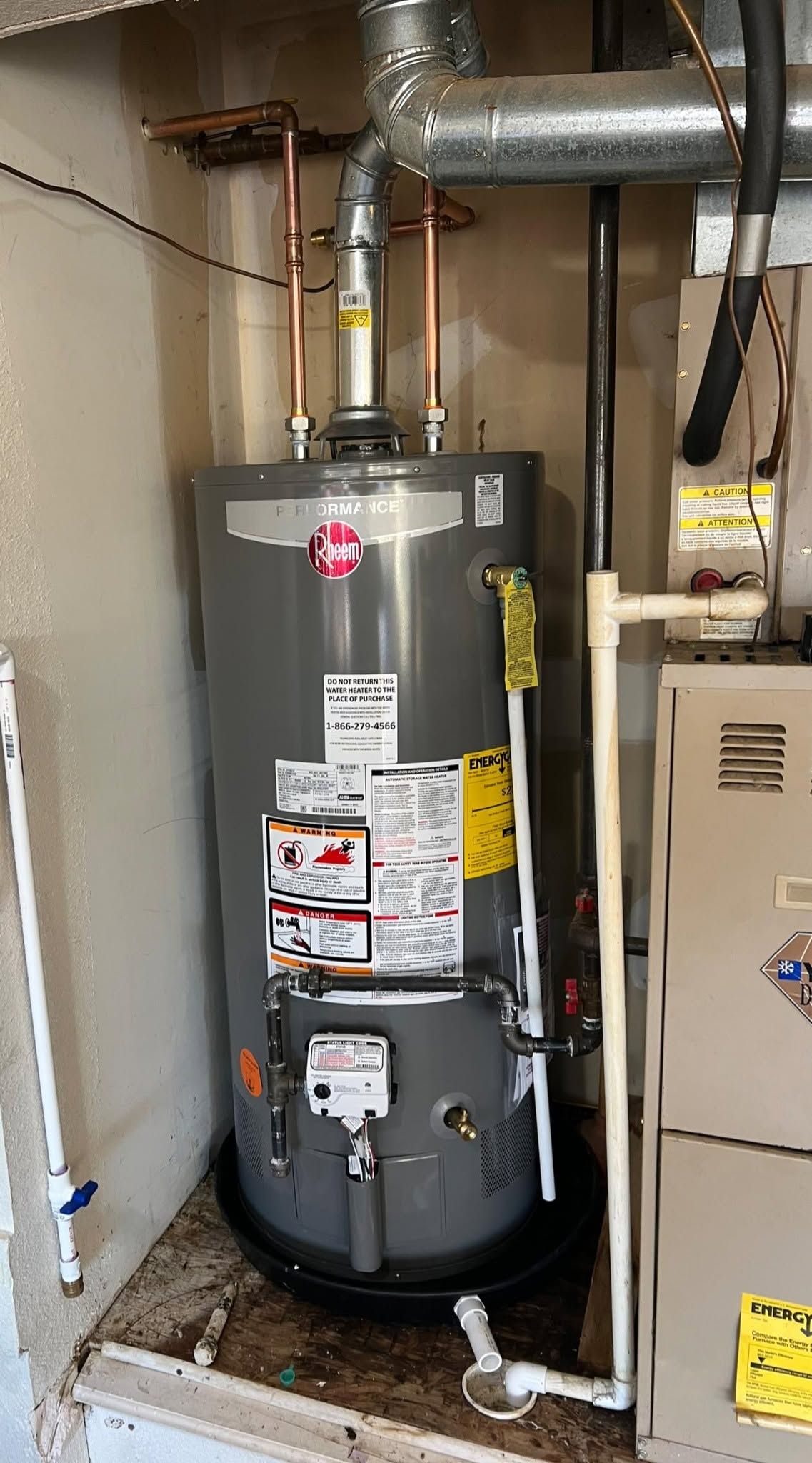 Gray water heater in a utility closet with pipes and a furnace beside it.