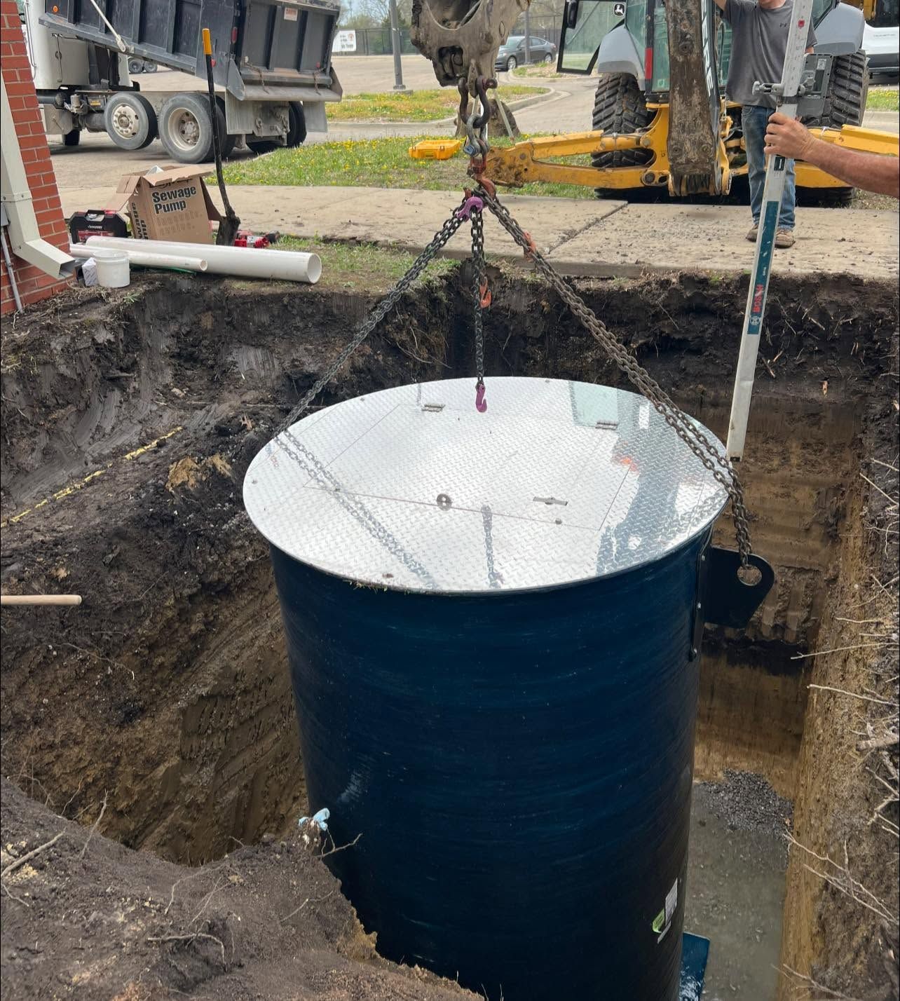 Excavator lifts a large blue steel cylinder from a muddy trench at a construction site