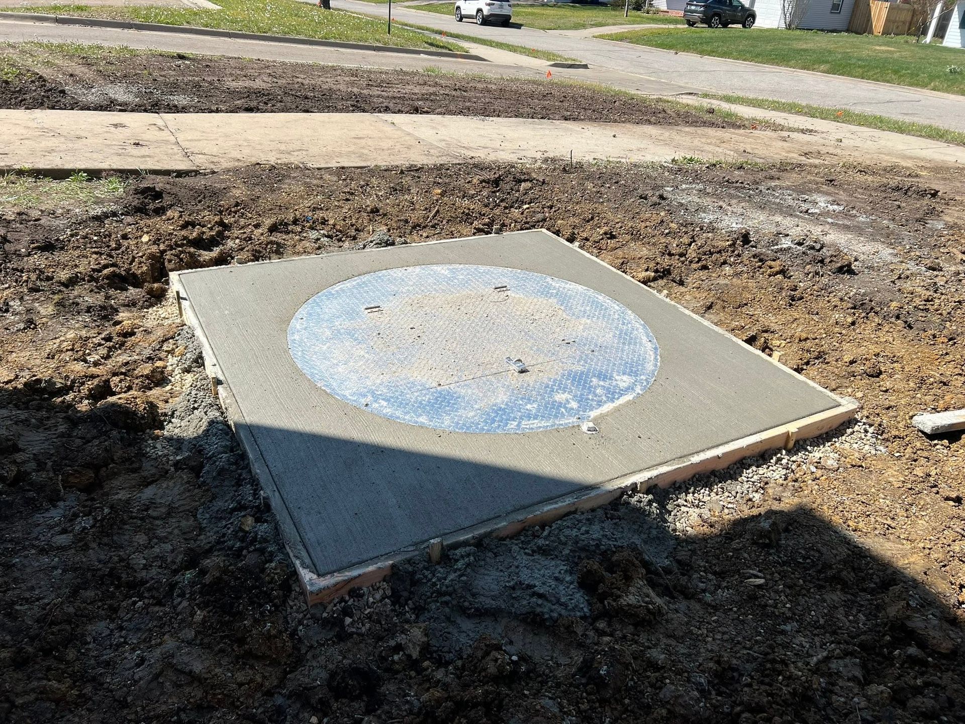 Concrete utility pad with a blue circular marking in a dirt-filled construction area by a sidewalk.