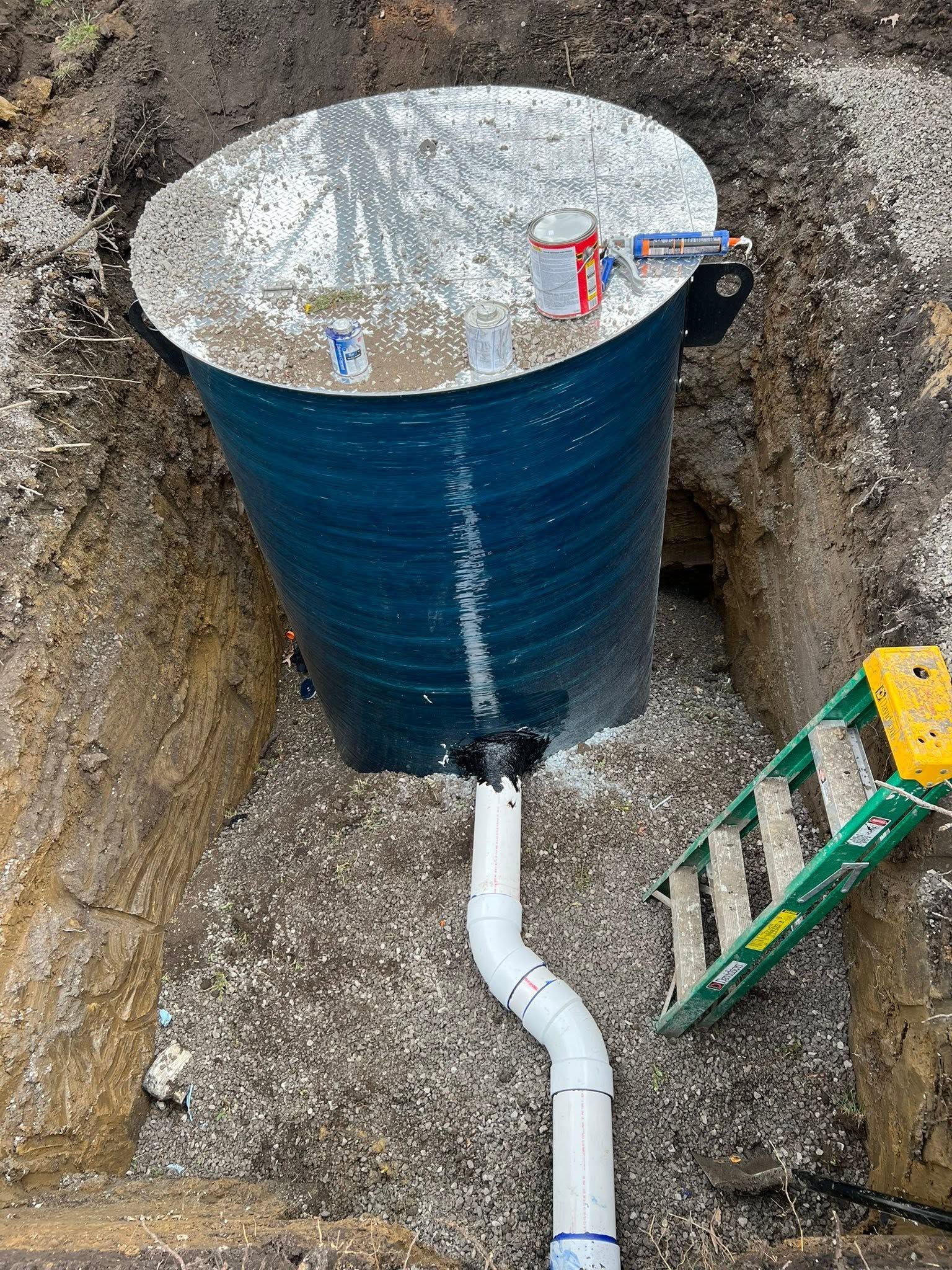 Blue water tank in a trench with a white pipe connected below it, beside a ladder and dirt walls