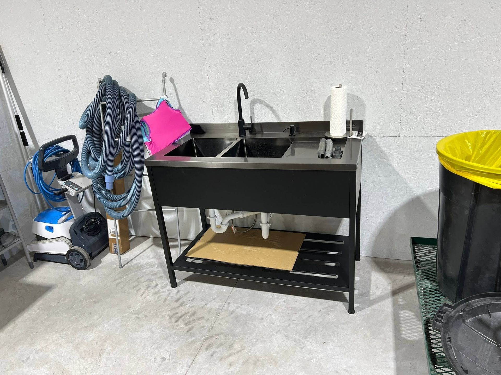 Utility sink and storage cabinet in a garage, with cleaning tools and a trash can nearby.