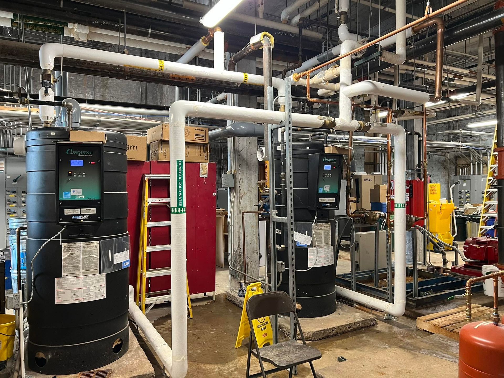 Industrial boiler room with large black water heaters, white pipes, and red equipment cabinets