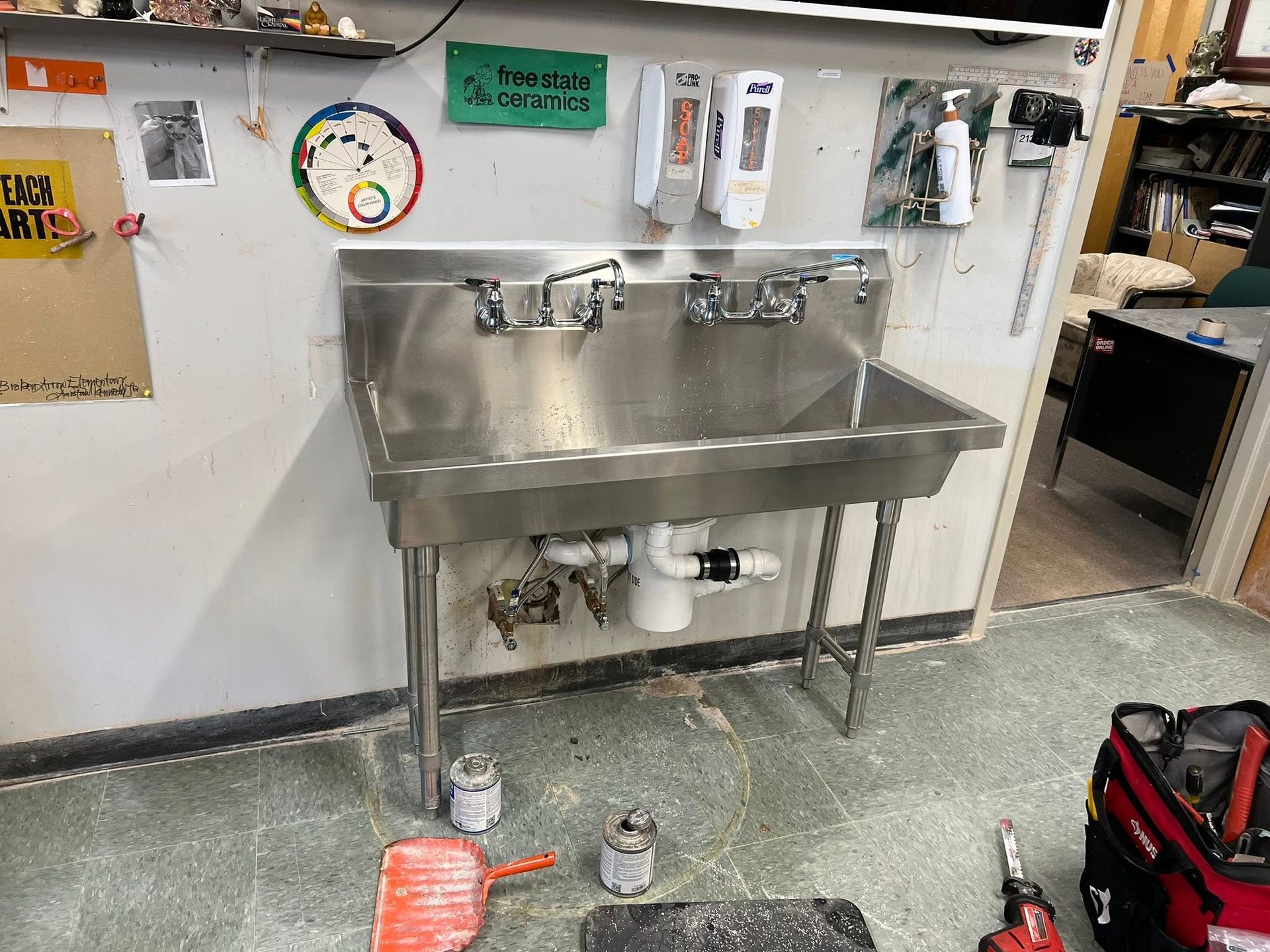 Stainless steel double sink in a utility room with cleaning supplies on the wall and a red tool bag on the floor