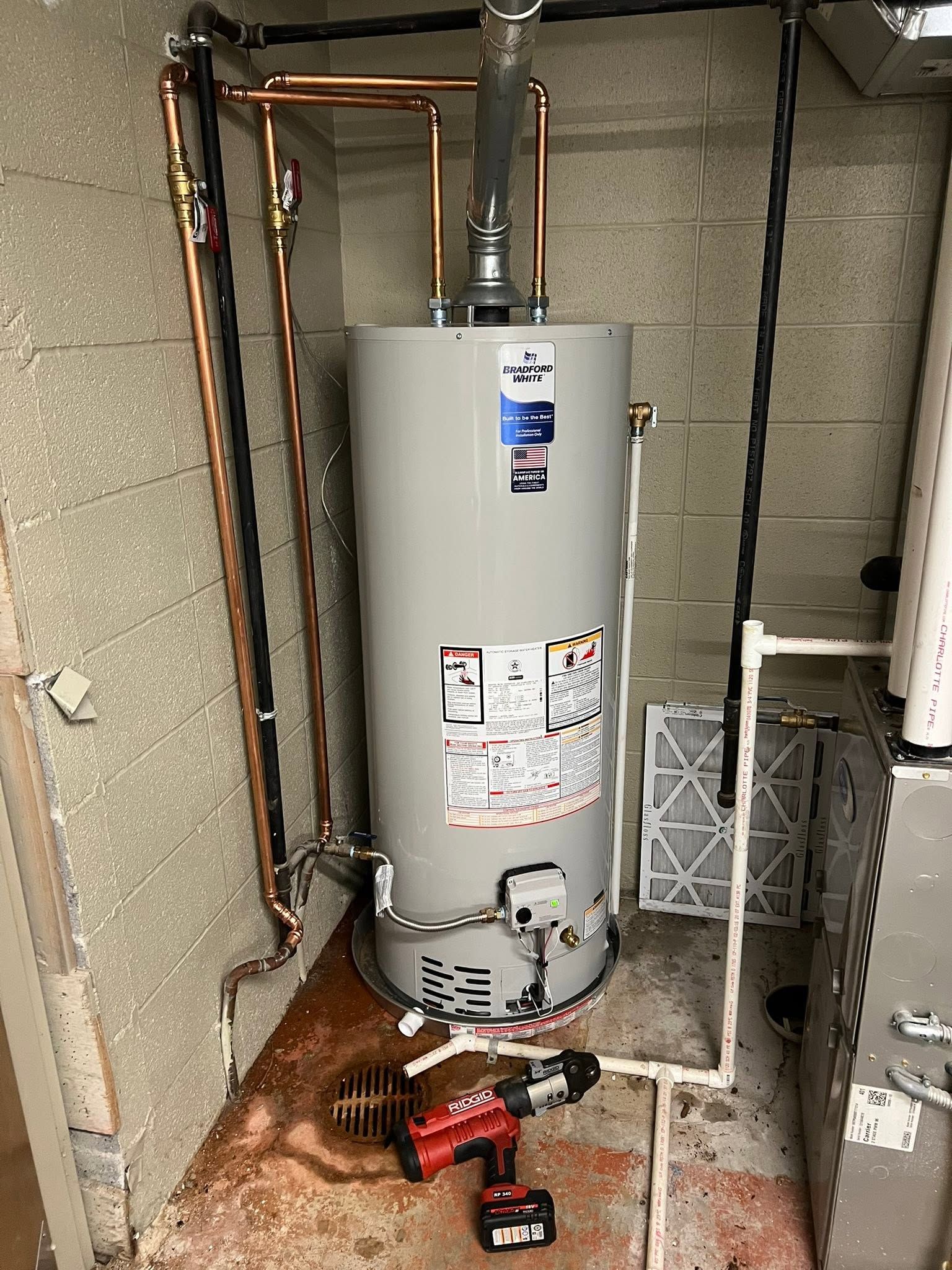 Water heater in a utility closet with exposed pipes and a red tool on the floor.