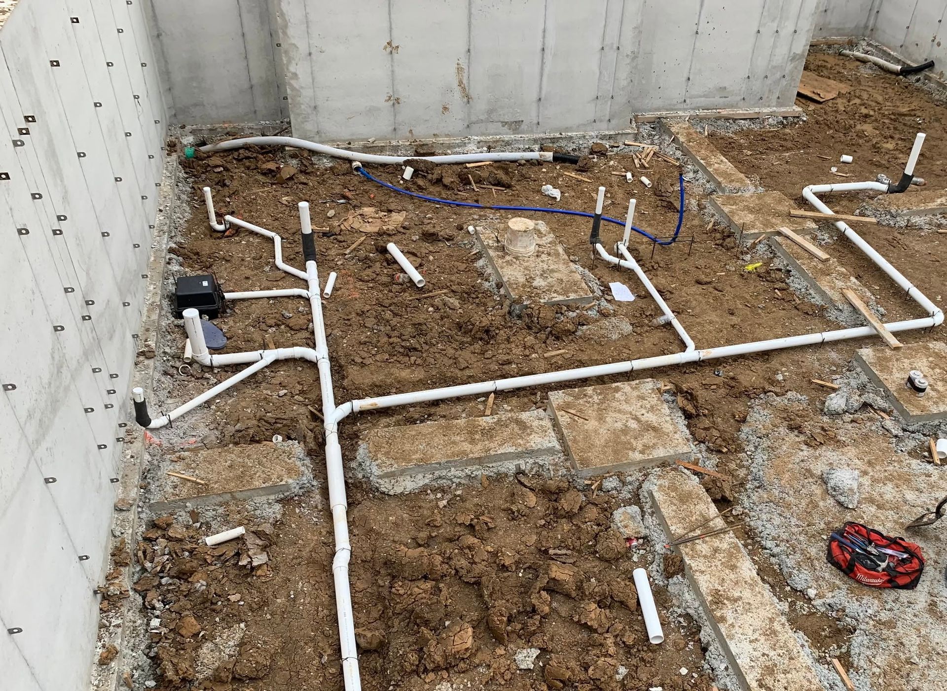 Plumbing pipes laid out in a dirt basement or crawl space beside concrete walls.