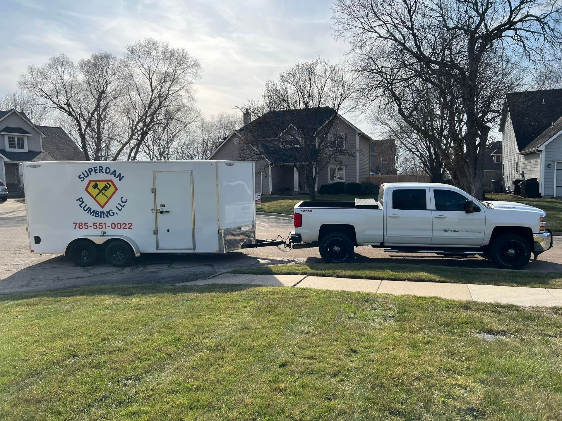 White pickup truck towing a white box trailer on a suburban street beside a grassy lawn.