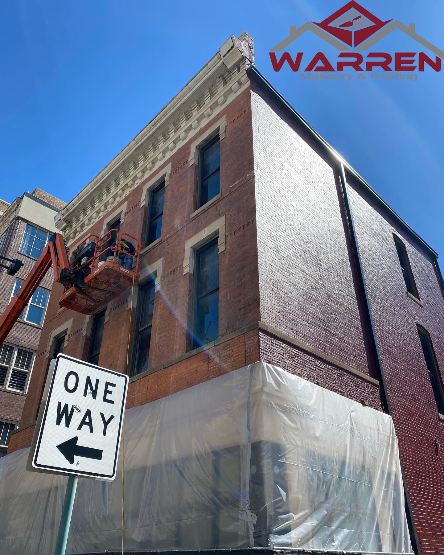 Brick corner building with fire escape, tarp-covered lower level, and a one-way sign against a blue sky