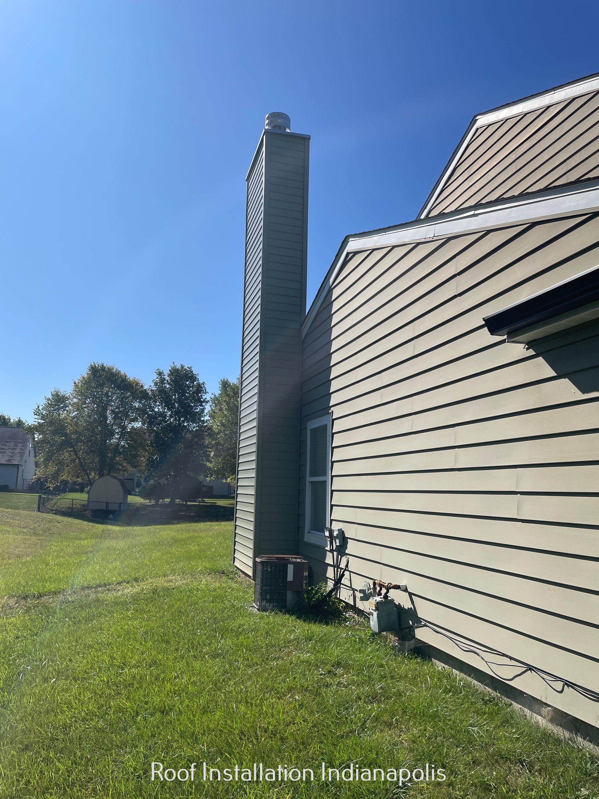 A side view of a house with tan siding, a tall chimney, an air conditioning unit, and a gas meter on a sunny lawn.